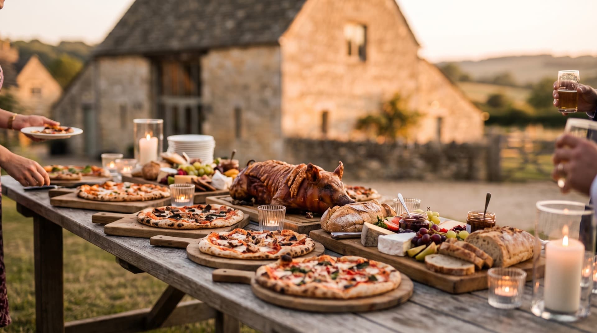 A feast table with wood-fired pizzas, a hog roast and grazing boards outside a Cotswold stone barn at golden hour