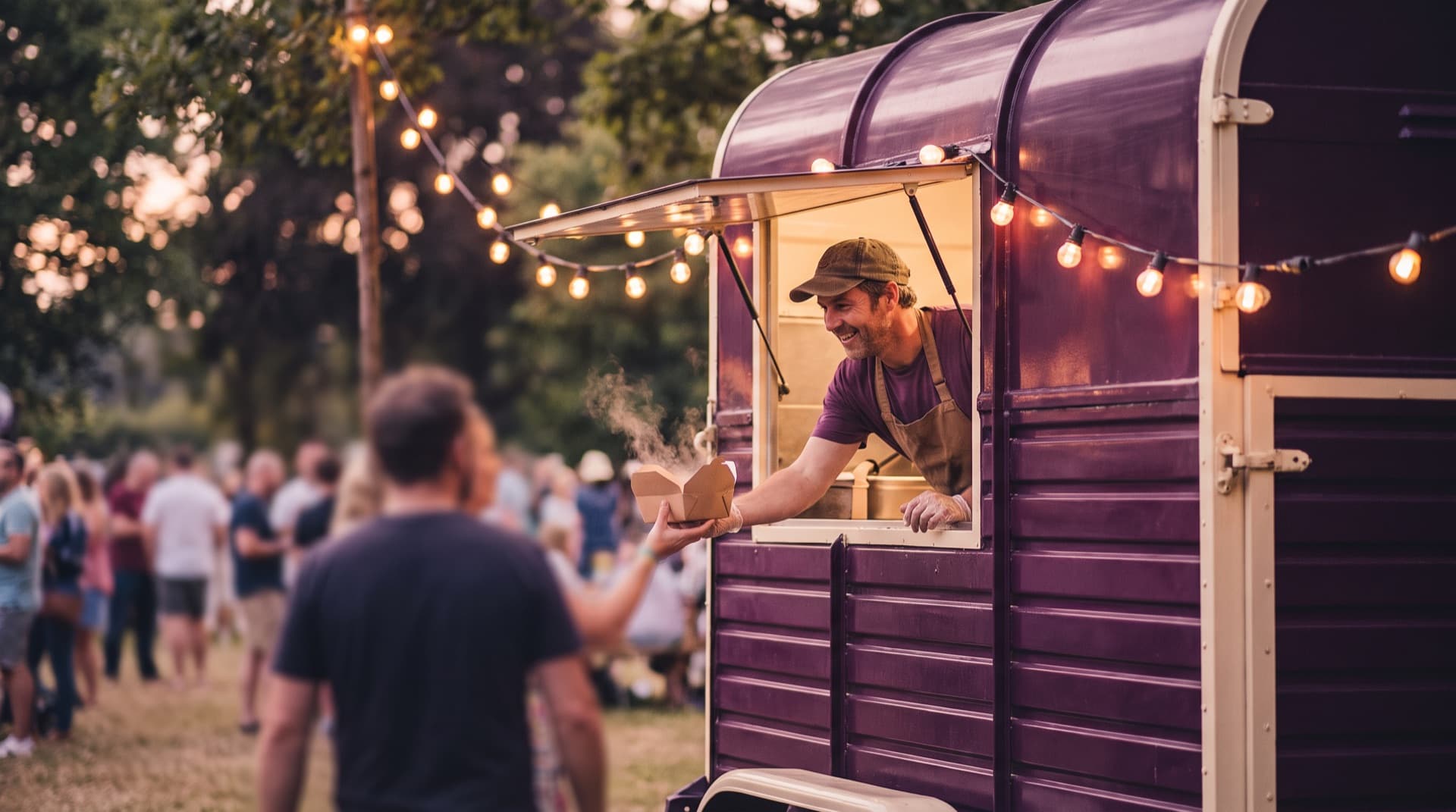 A street food trader serving from a horsebox van under festoon lights at dusk